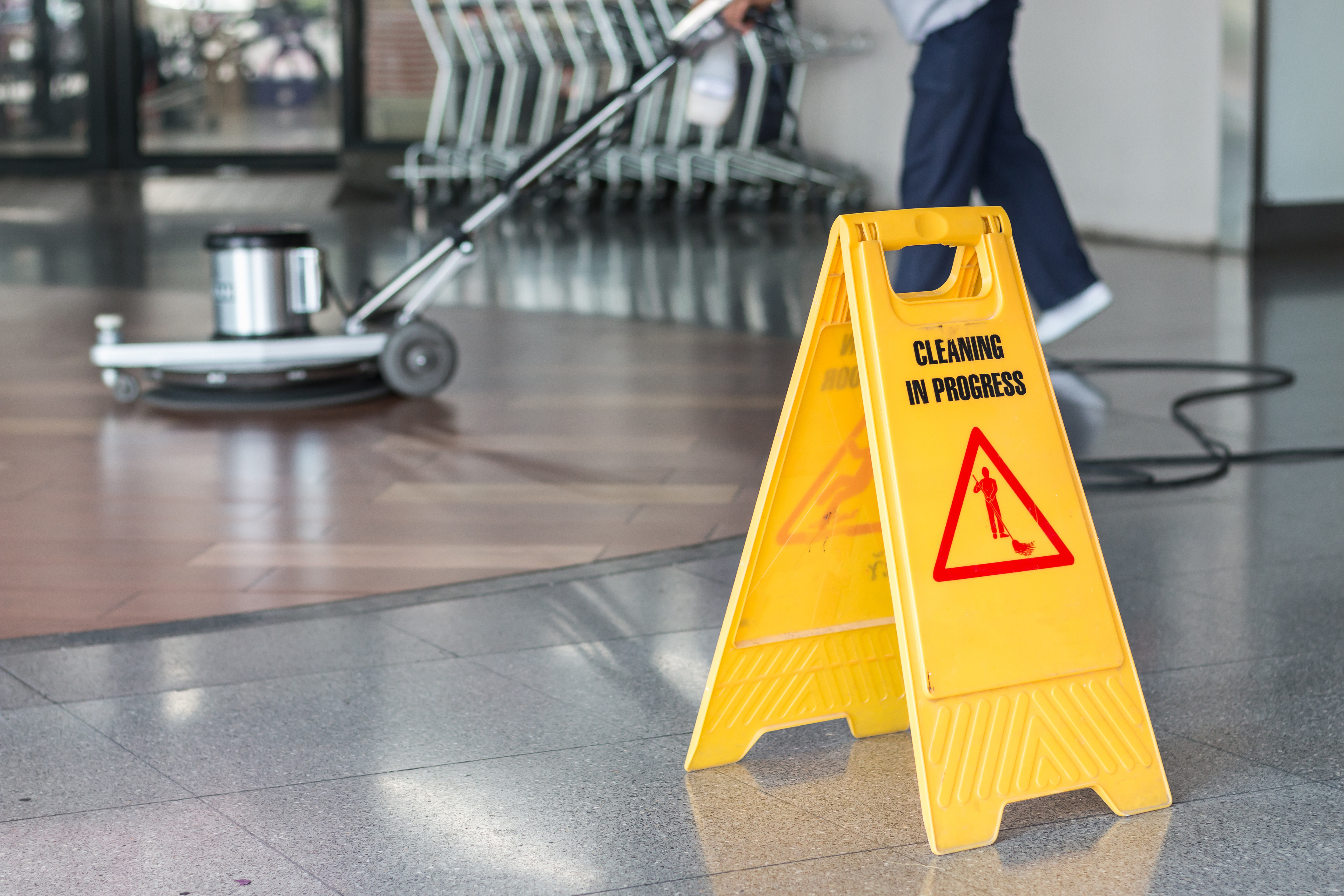 Yellow wet floor sign with "CLEANING IN PROGRESS" text and warning symbol on polished floor with cleaning equipment in background
