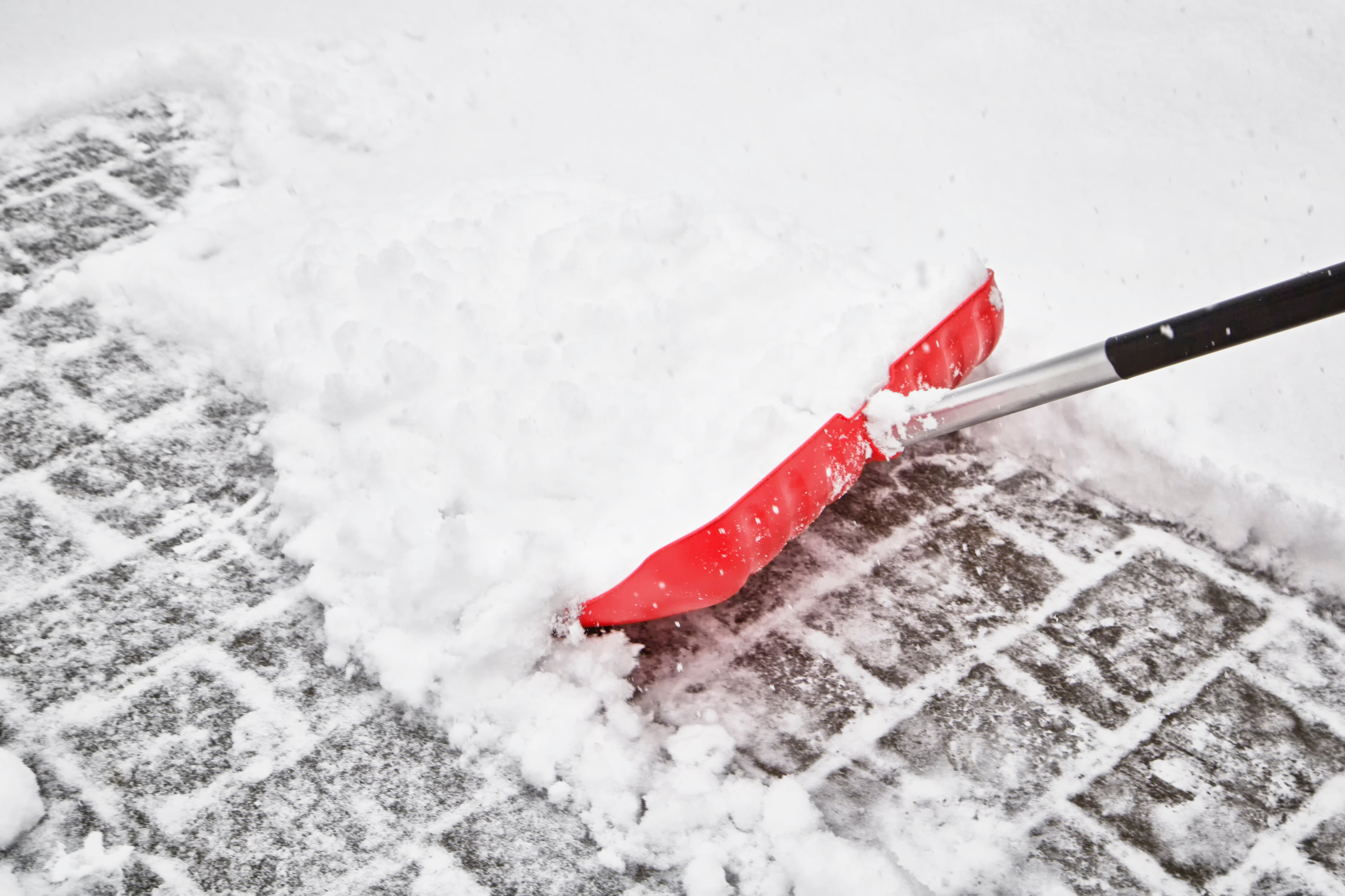 Red snow shovel clearing snow and ice from brick pavement