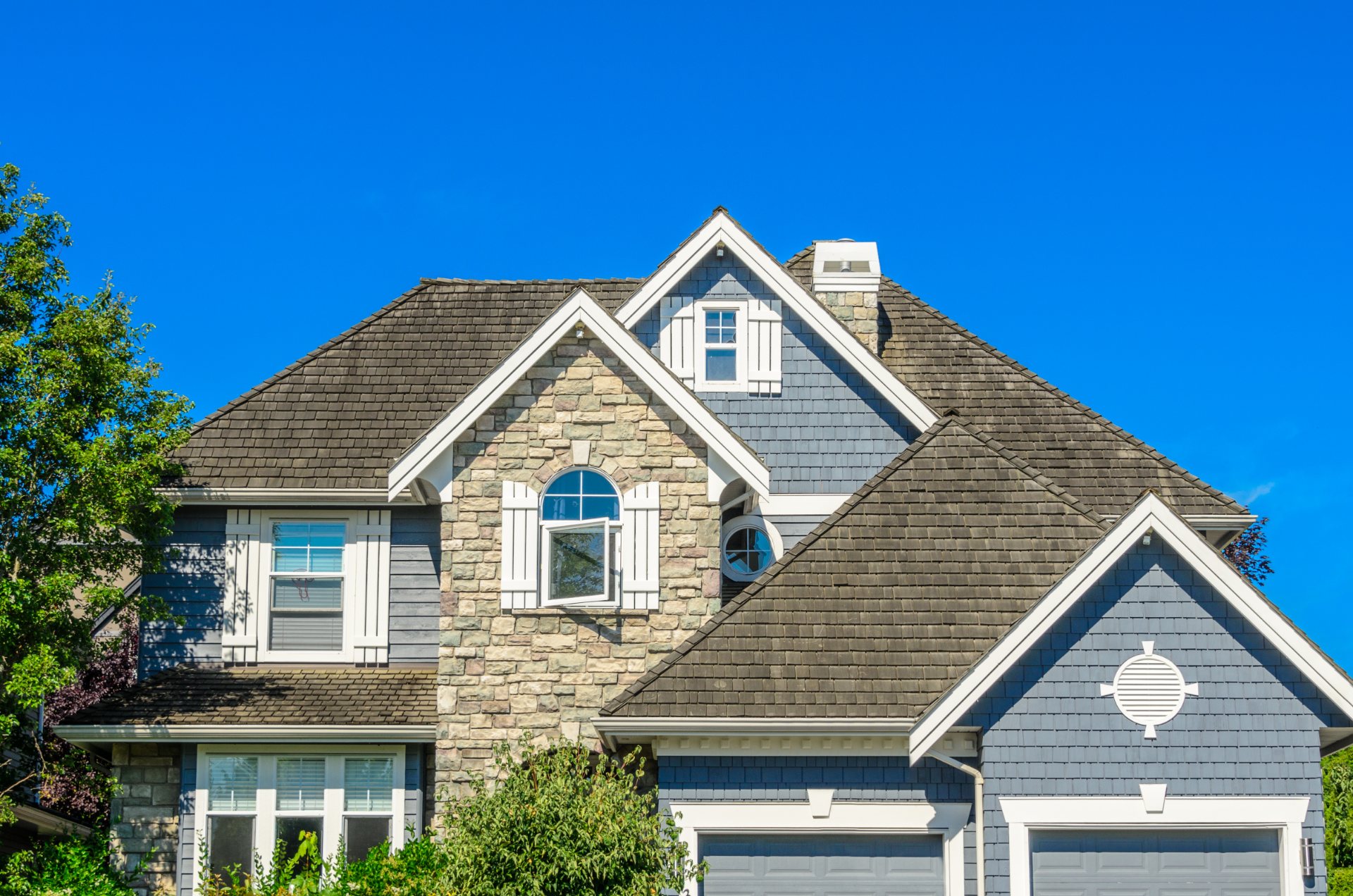 Two-story stone and blue siding home with peaked roofline and two-car garage under clear blue sky