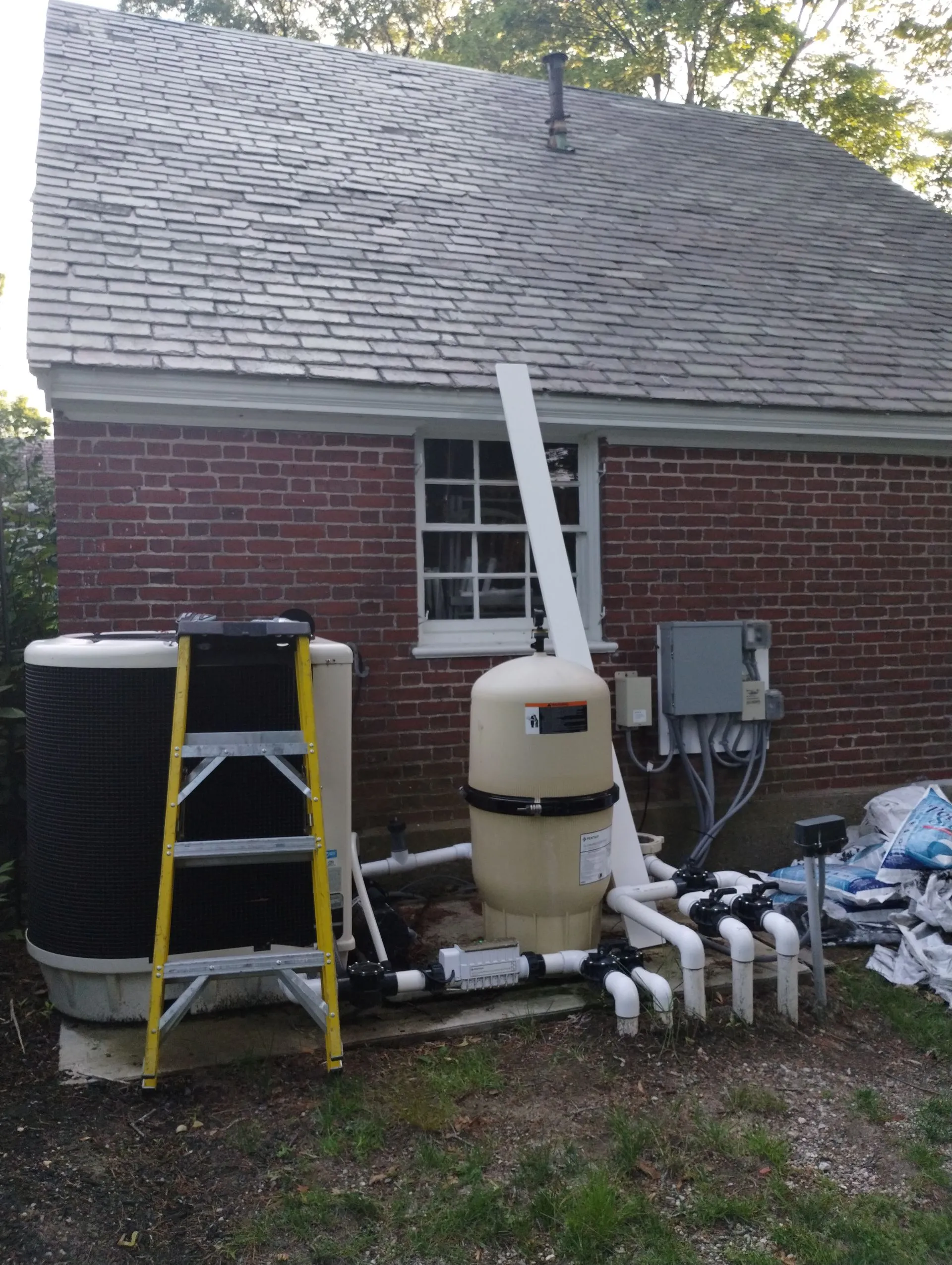 Residential pool equipment installation showing tanks, pipes, and filtration system against brick house
