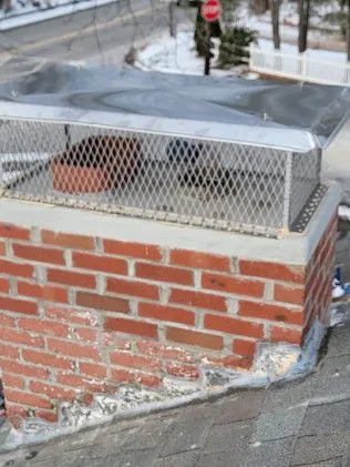 Metal chimney cap with chain-link mesh on red brick chimney in snowy setting