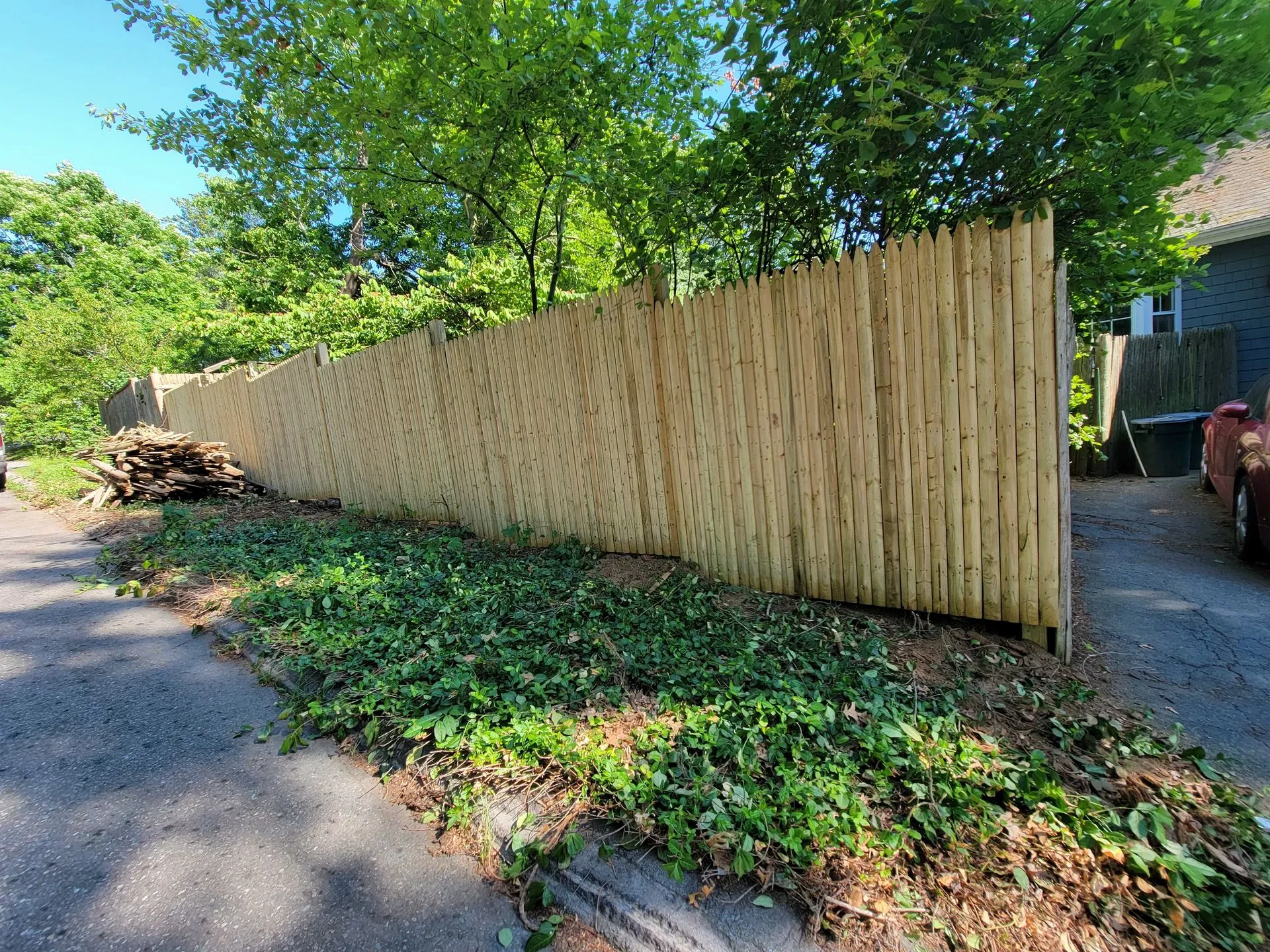 Wooden fence along residential driveway with overgrown vegetation and debris at base