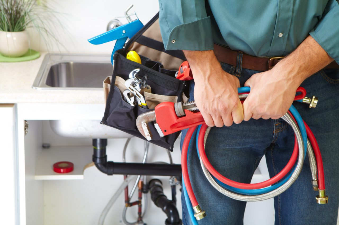 Plumber holding pipe wrench and coiled hoses in kitchen with sink and toolbelt